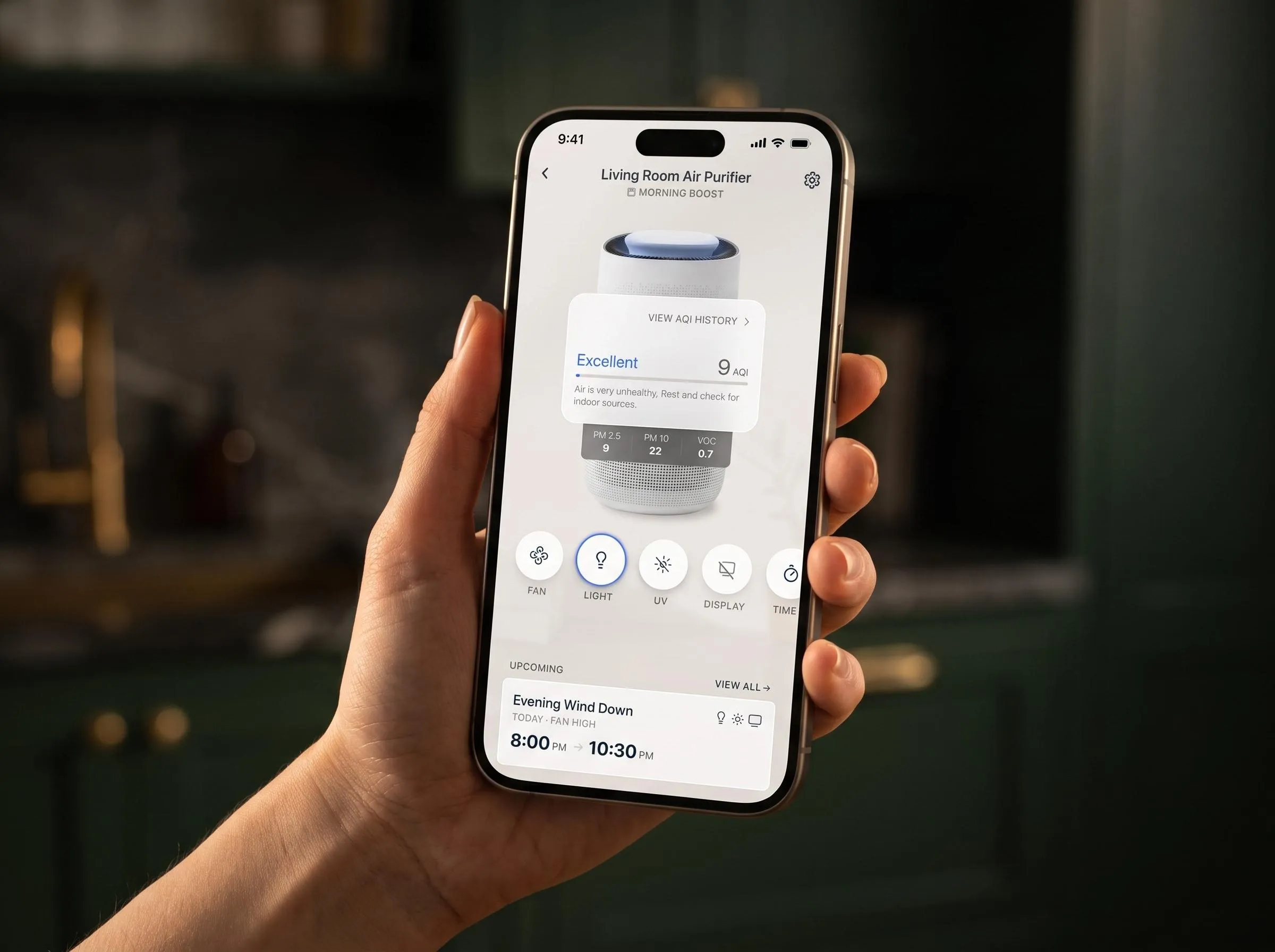 A hand holding a phone showing the air purifier screen. Fan, light, UV, display and timer sit in one row of round buttons beneath the device, above the next scheduled run.